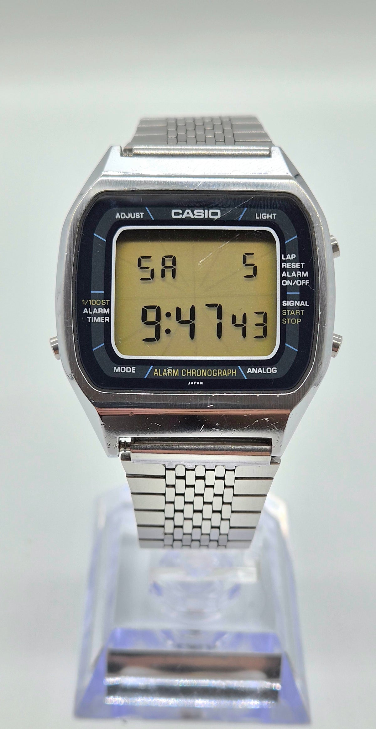 A classic Casio digital wrist watch with a silver metal case and link bracelet, featuring a black bezel and a yellow-green LCD display showing the time 9:47:43. The watch is displayed on a clear acrylic stand against a light gray background.