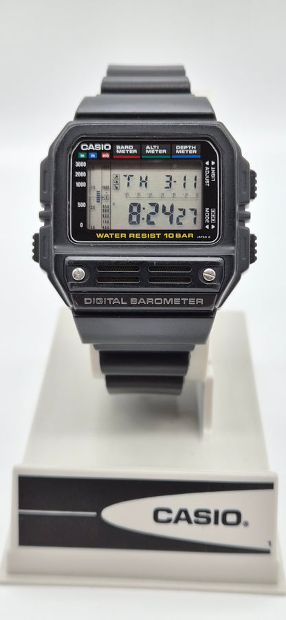 A Casio digital barometer watch with a black rugged case and strap, featuring a digital LCD display, shown resting on a white and black Casio display stand against a light gray background.