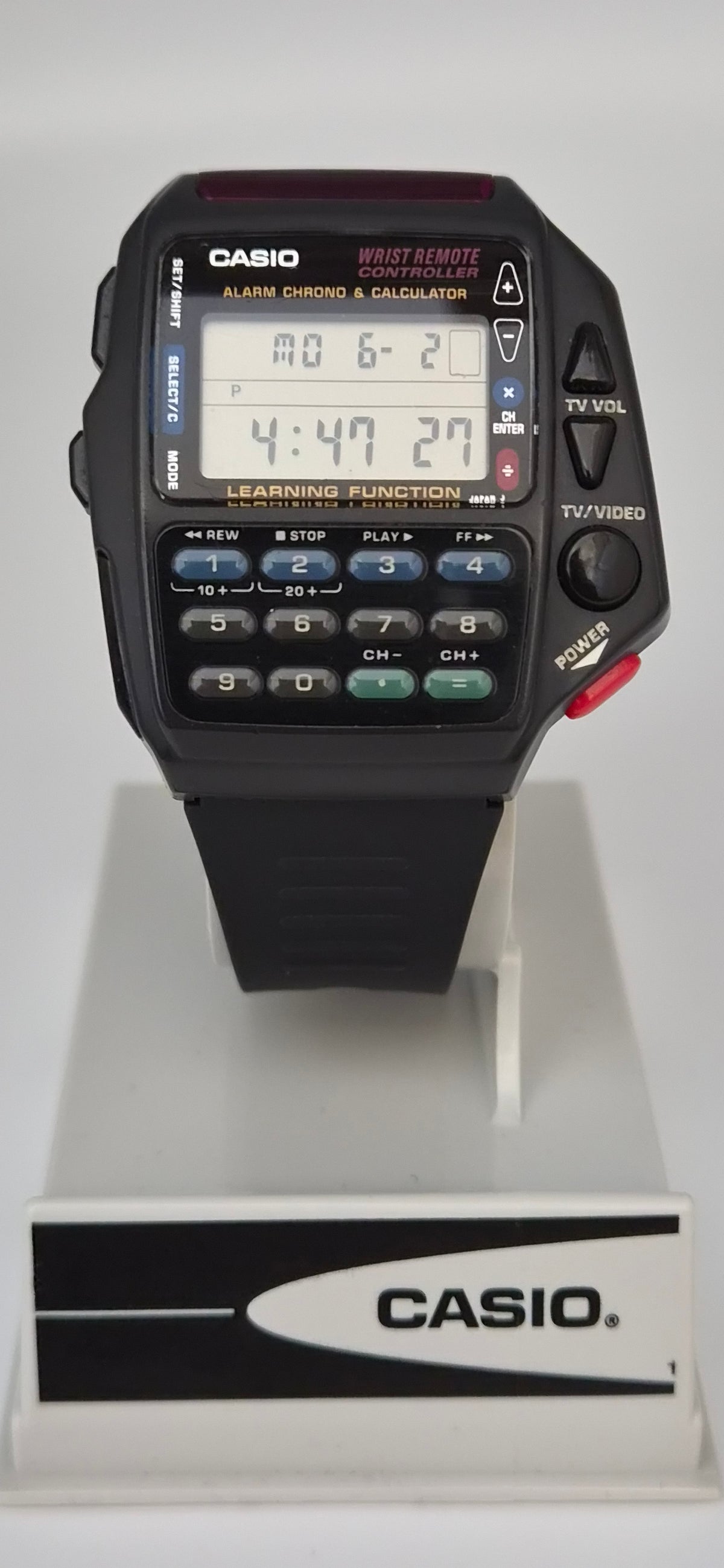 A Casio digital alarm clock watch with a black strap and face, featuring a digital display, numeric keypad, and red power button, shown on a white display stand with a Casio logo plaque.
