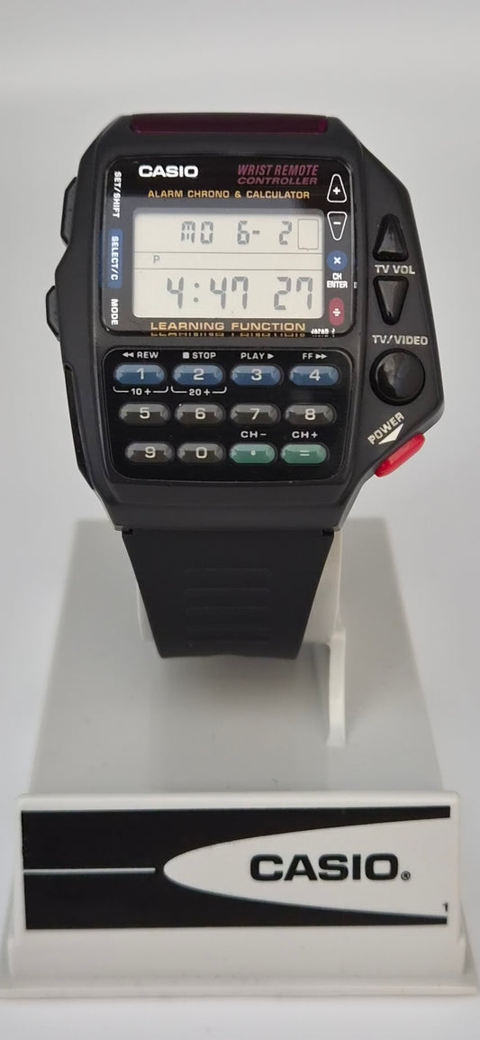 A Casio digital alarm clock watch with a black strap and face, featuring a digital display, numeric keypad, and red power button, shown on a white display stand with a Casio logo plaque.