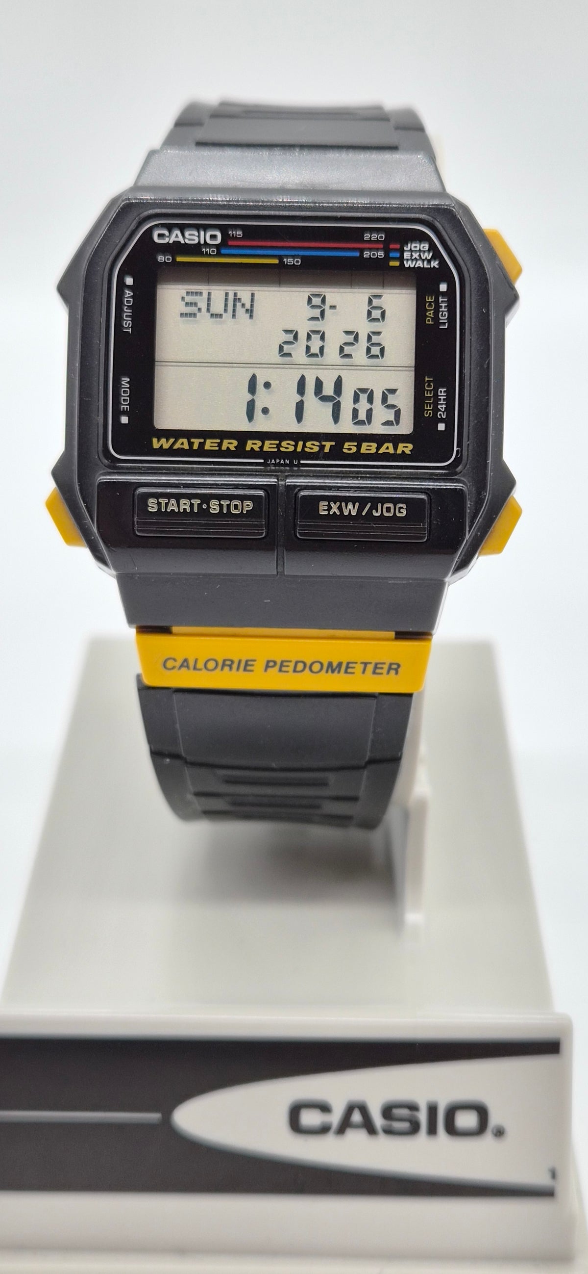 A Casio digital wrist watch with a black and gray case and strap, featuring yellow accents on the side buttons and a yellow band section. The watch is displayed on a white stand with a Casio logo at the bottom.