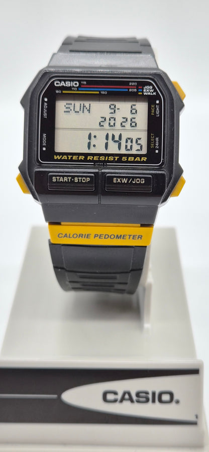 A Casio digital wrist watch with a black and gray case and strap, featuring yellow accents on the side buttons and a yellow band section. The watch is displayed on a white stand with a Casio logo at the bottom.