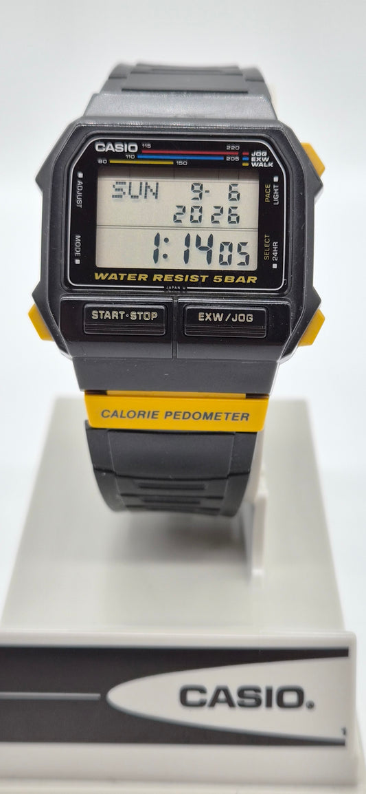 A Casio digital wrist watch with a black and gray case and strap, featuring yellow accents on the side buttons and a yellow band section. The watch is displayed on a white stand with a Casio logo at the bottom.