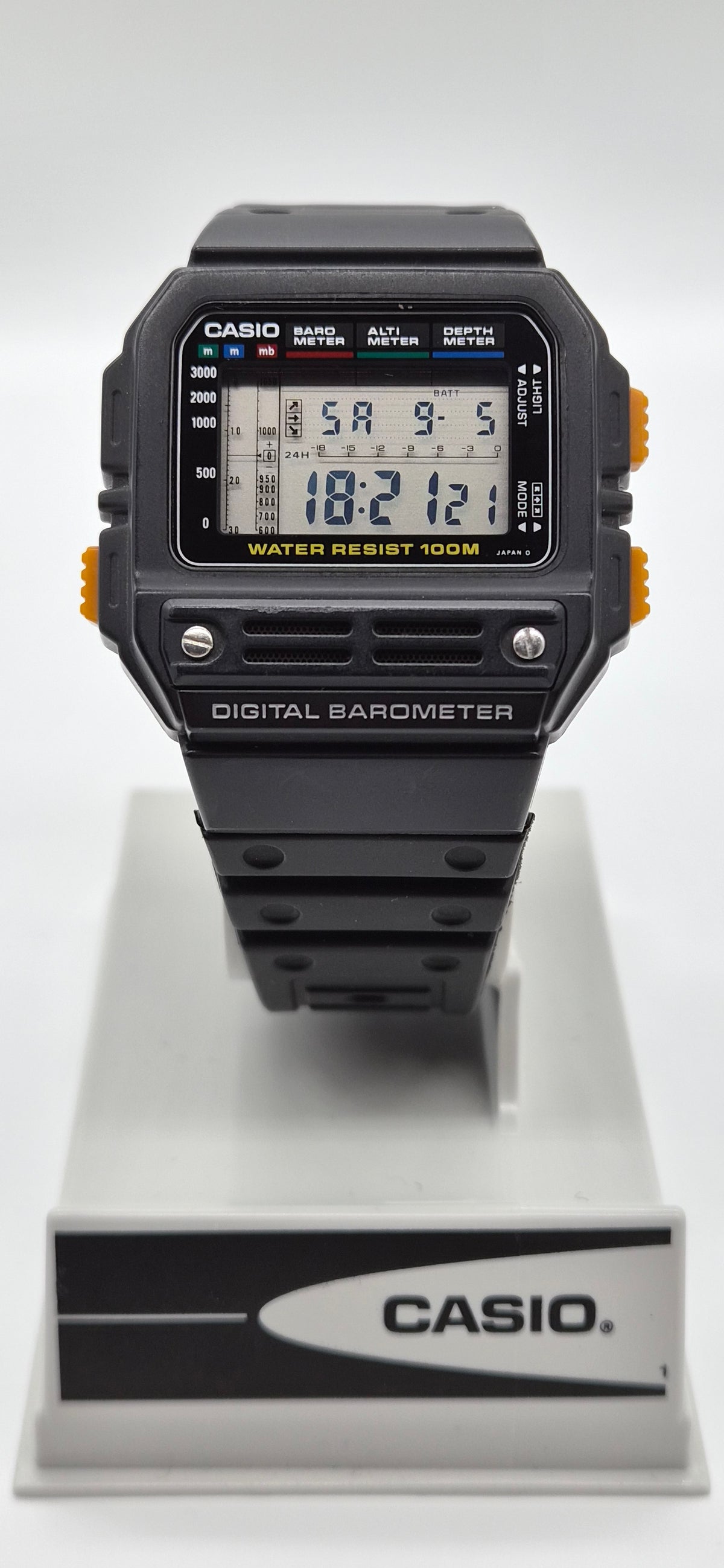 A Casio digital barometer watch with a black strap and case, featuring orange side accents and a digital LCD display, shown resting on a white and gray Casio display stand against a white background.