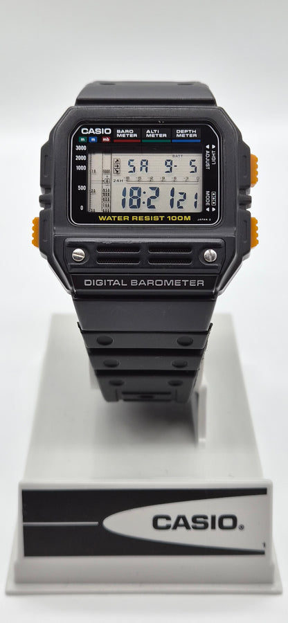 A Casio digital barometer watch with a black strap and case, featuring orange side accents and a digital LCD display, shown resting on a white and gray Casio display stand against a white background.
