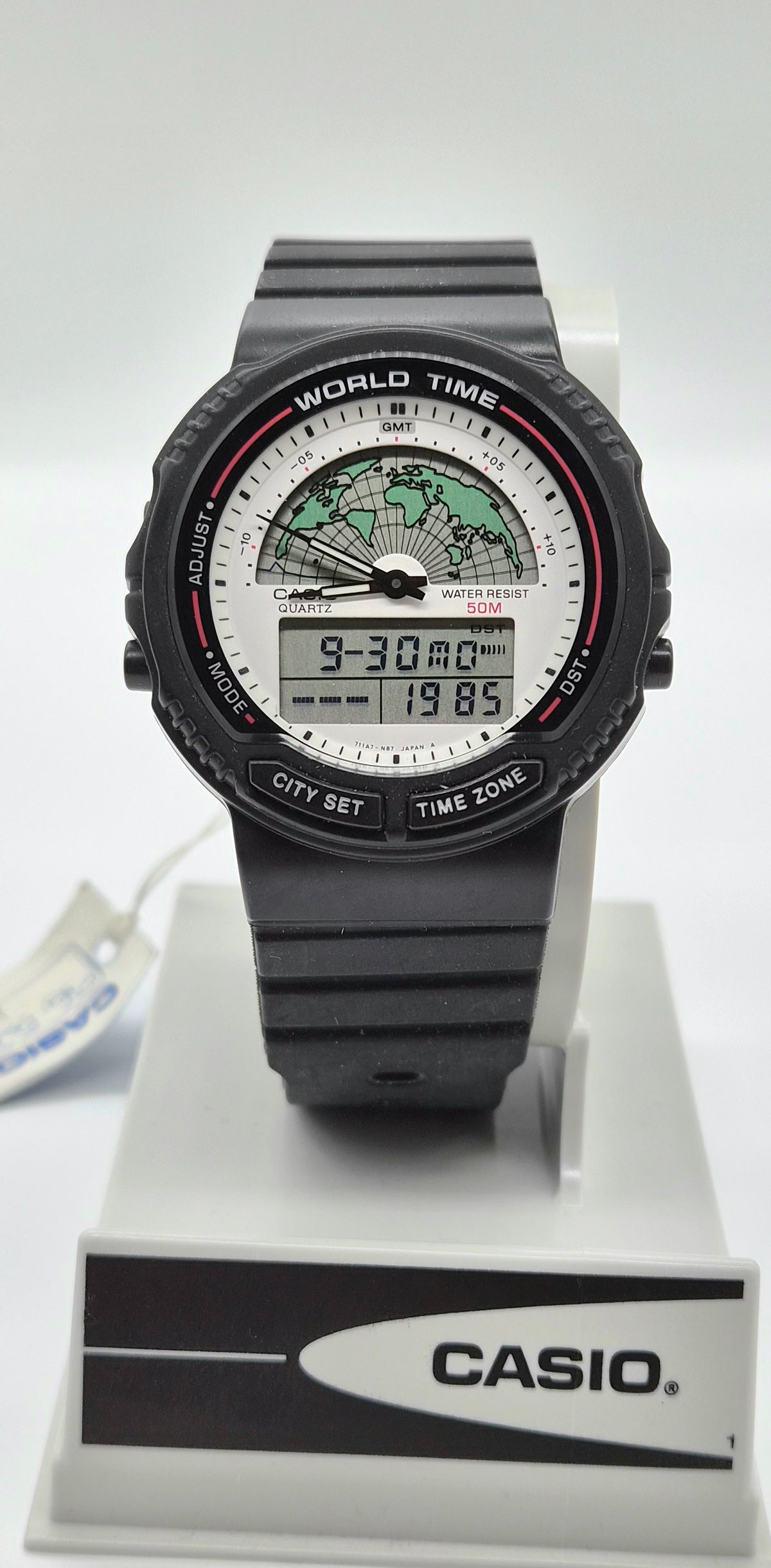 A Casio World Time digital watch with a black strap and case, featuring a white dial with a green world map graphic and digital time display, shown on a white display stand with a Casio logo.