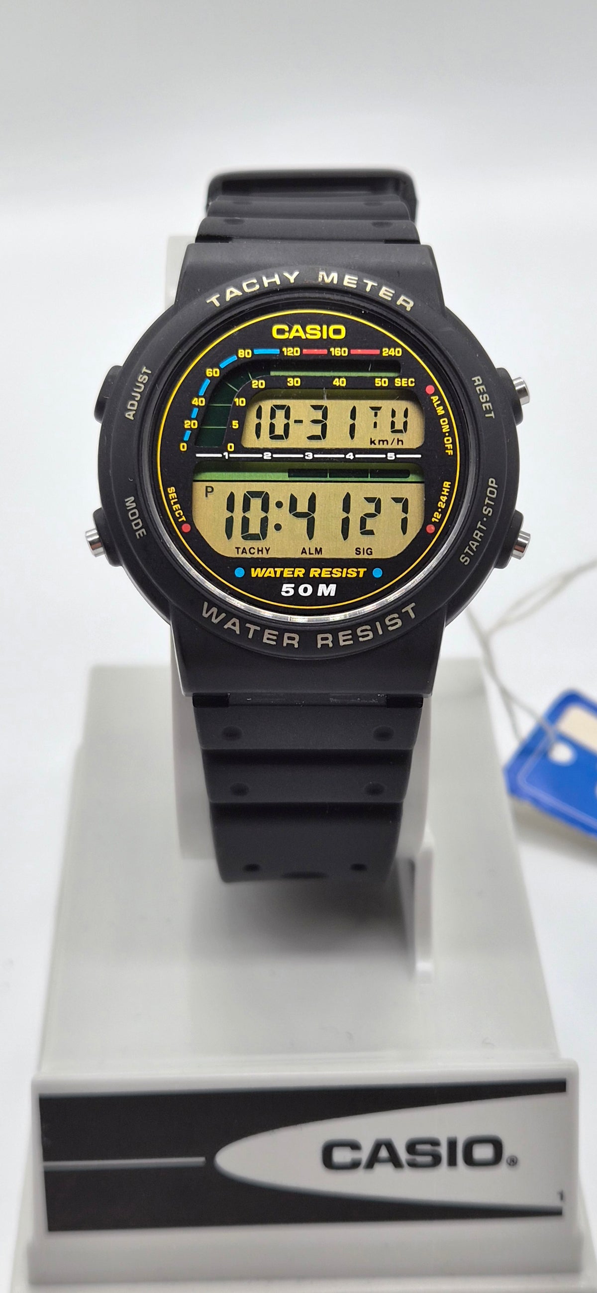 A Casio digital watch with a black resin strap and case, featuring a yellow and black analog-style dial display, resting on a white Casio display stand.