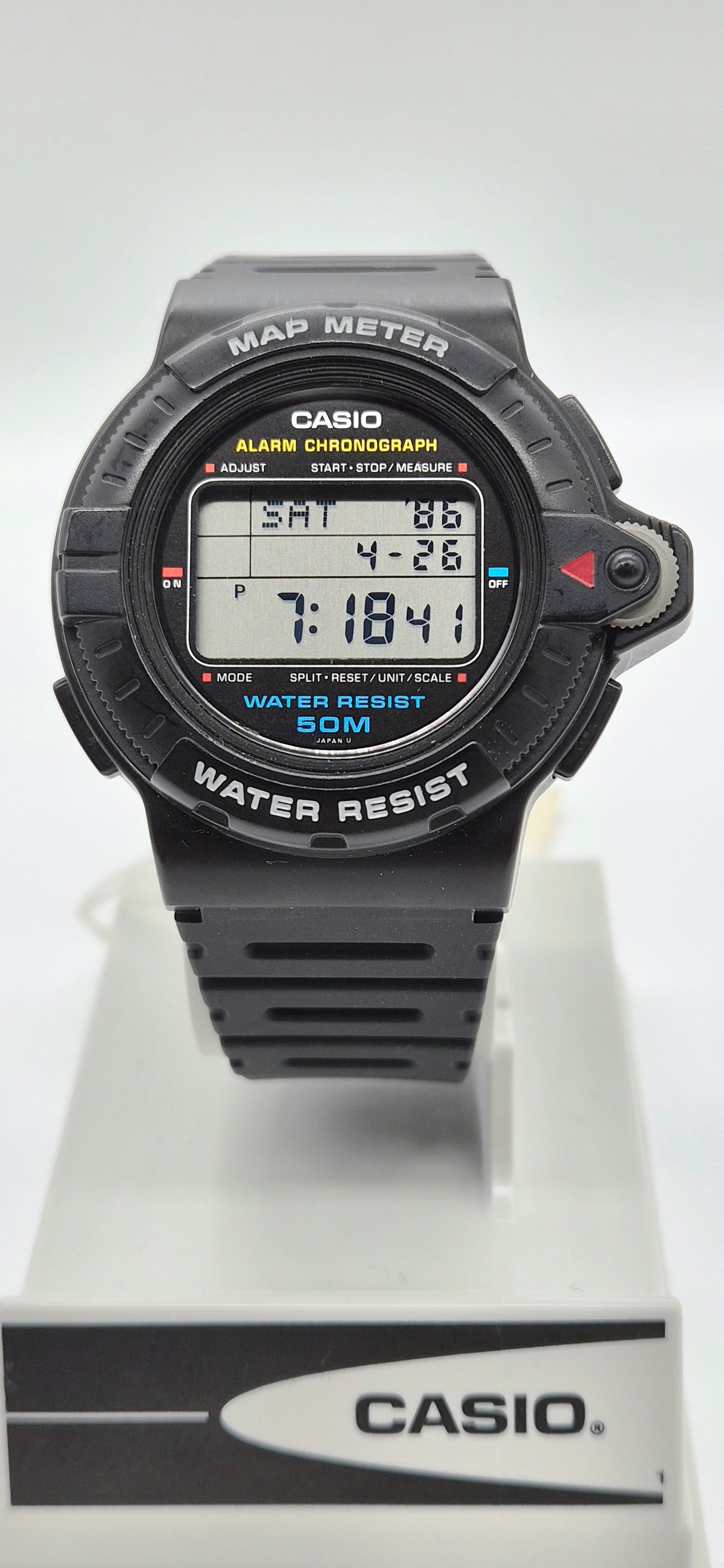 A Casio digital map and compass watch with a black strap and case, displayed on a white stand with a branded base against a white background.