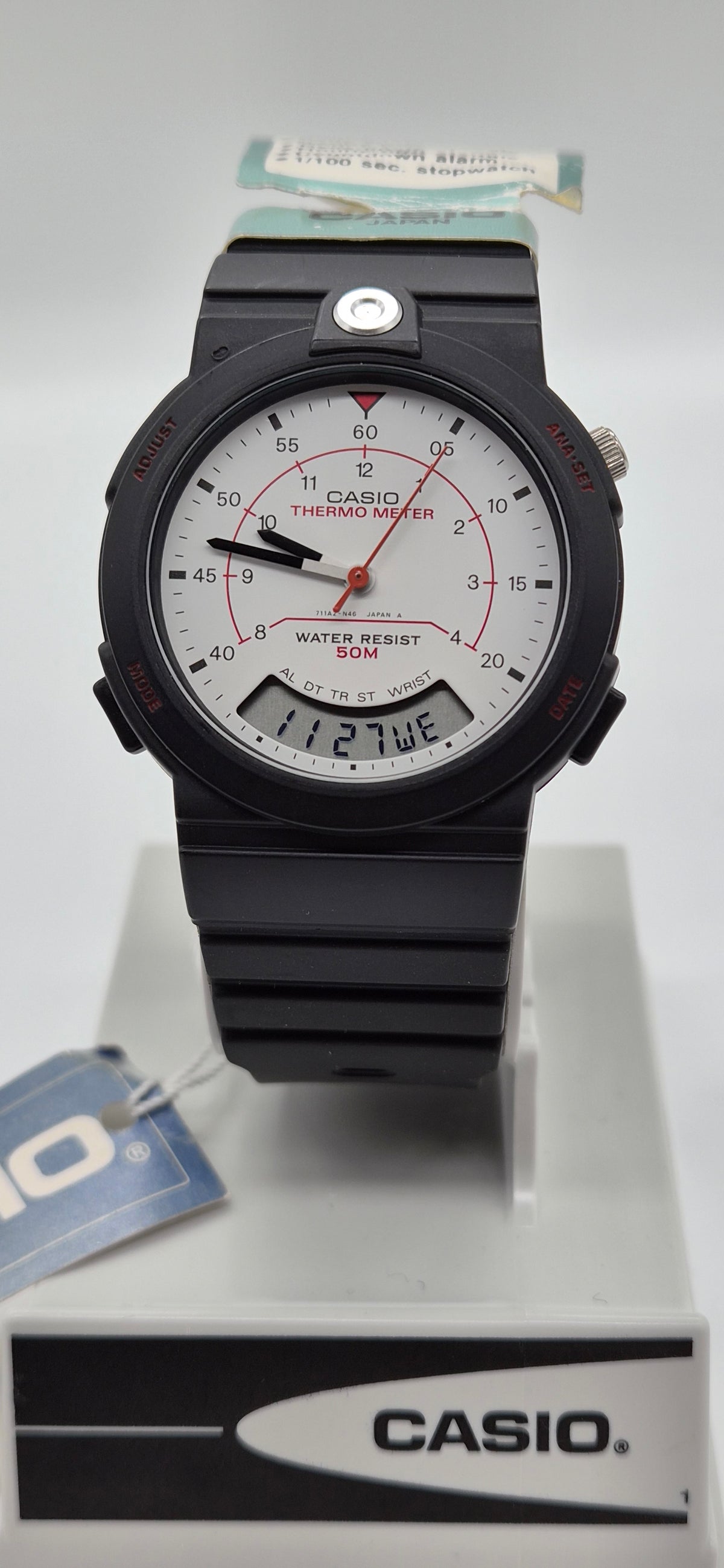 A Casio analog watch with a black resin case and strap, featuring a white dial with red accents and a digital day window, displayed on a white stand with a Casio-branded box.