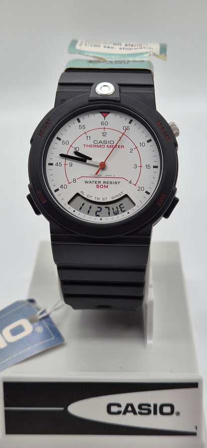 A Casio analog watch with a black resin case and strap, featuring a white dial with red accents and a digital day window, displayed on a white stand with a Casio-branded box.