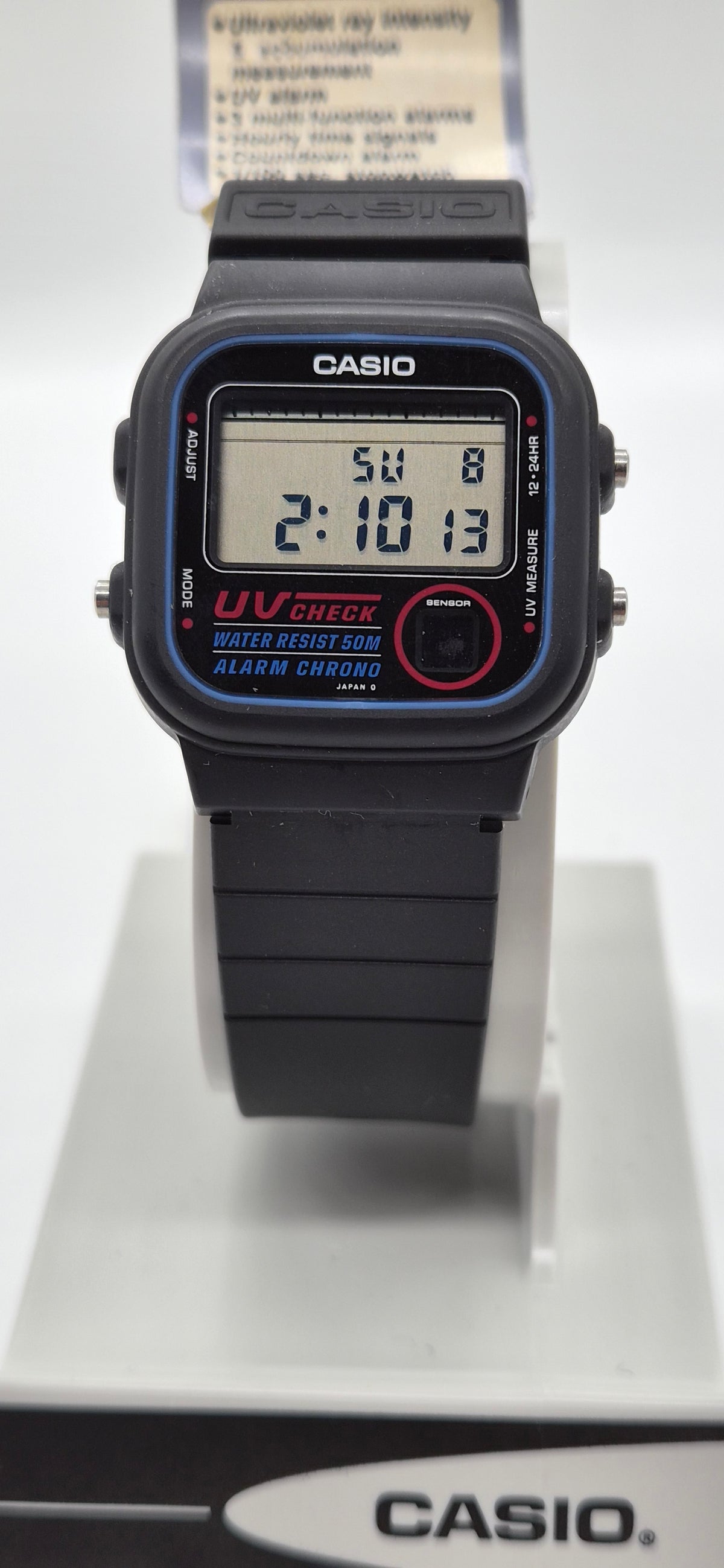 A Casio digital wrist watch with a black strap and case, featuring blue bezel detailing, a red circular button, and a digital display showing the time 2:10:13.