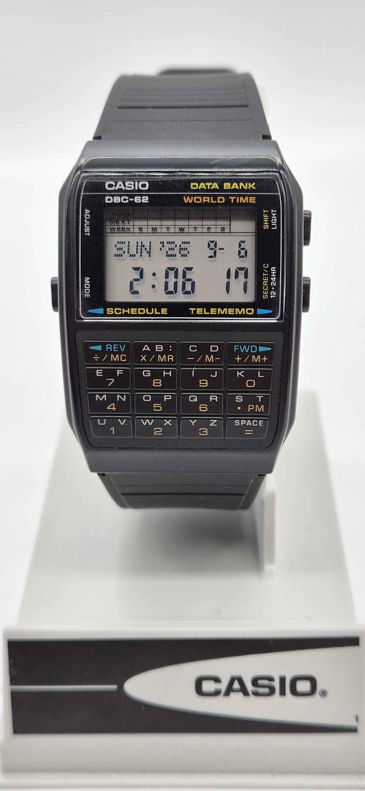 A Casio DCG-82 World Time digital calculator watch with a black plastic strap and case, featuring a yellow display screen and black numeric keypad, displayed on a white stand with a branded base.