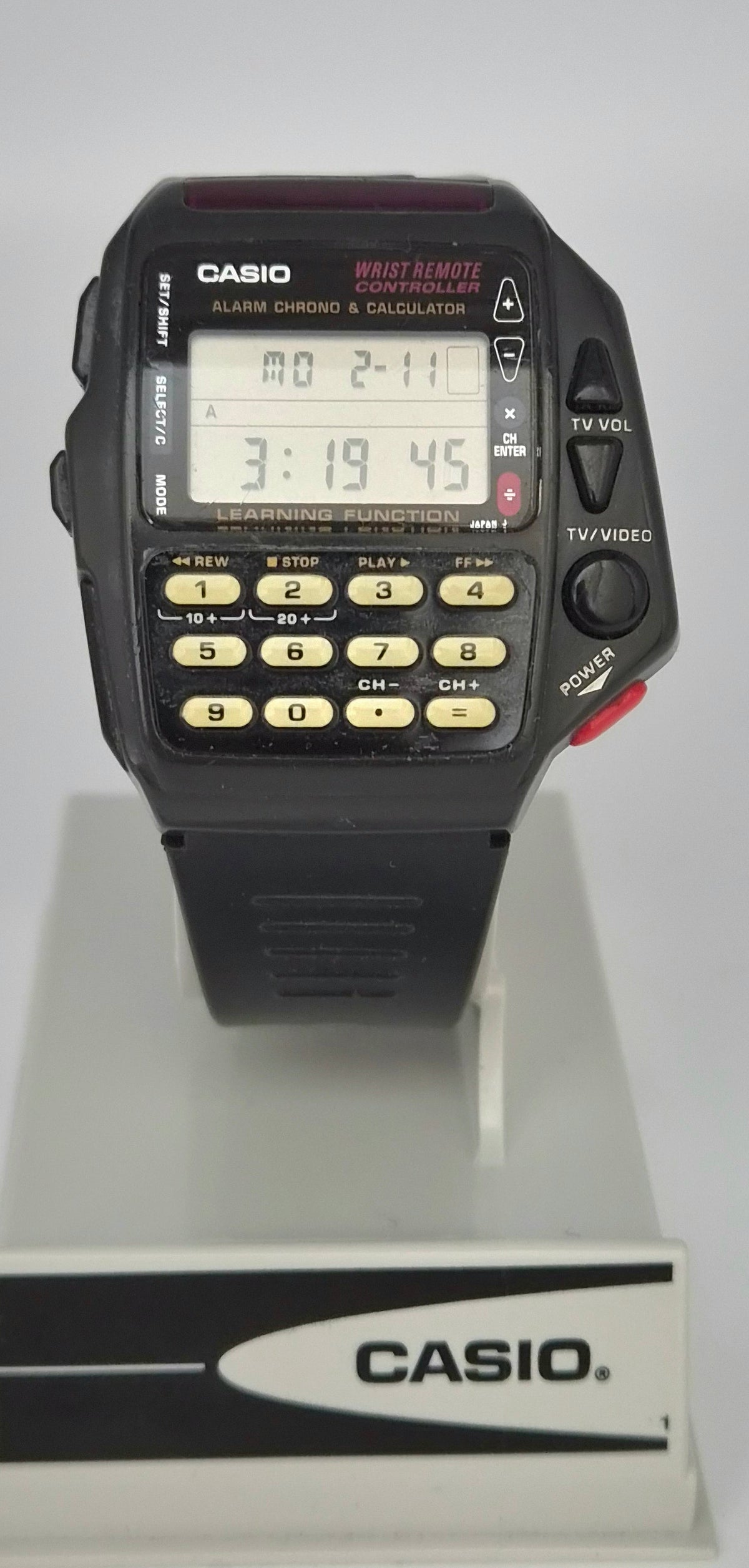 A vintage Casio wrist watch with a black plastic case, digital LCD display, and numeric keypad, shown resting on a white display stand with a Casio logo.