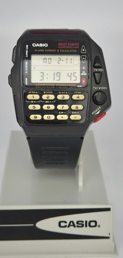 A vintage Casio wrist watch with a black plastic case, digital LCD display, and numeric keypad, shown resting on a white display stand with a Casio logo.