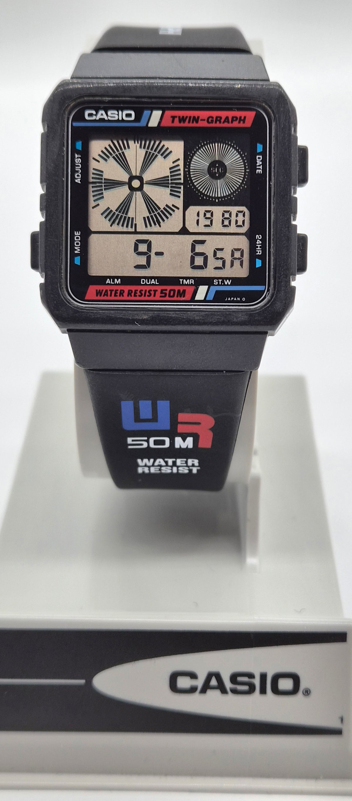 A Casio dual display watch with a black resin case and strap, featuring analog and digital screens, shown on a white display stand with a Casio-branded base.