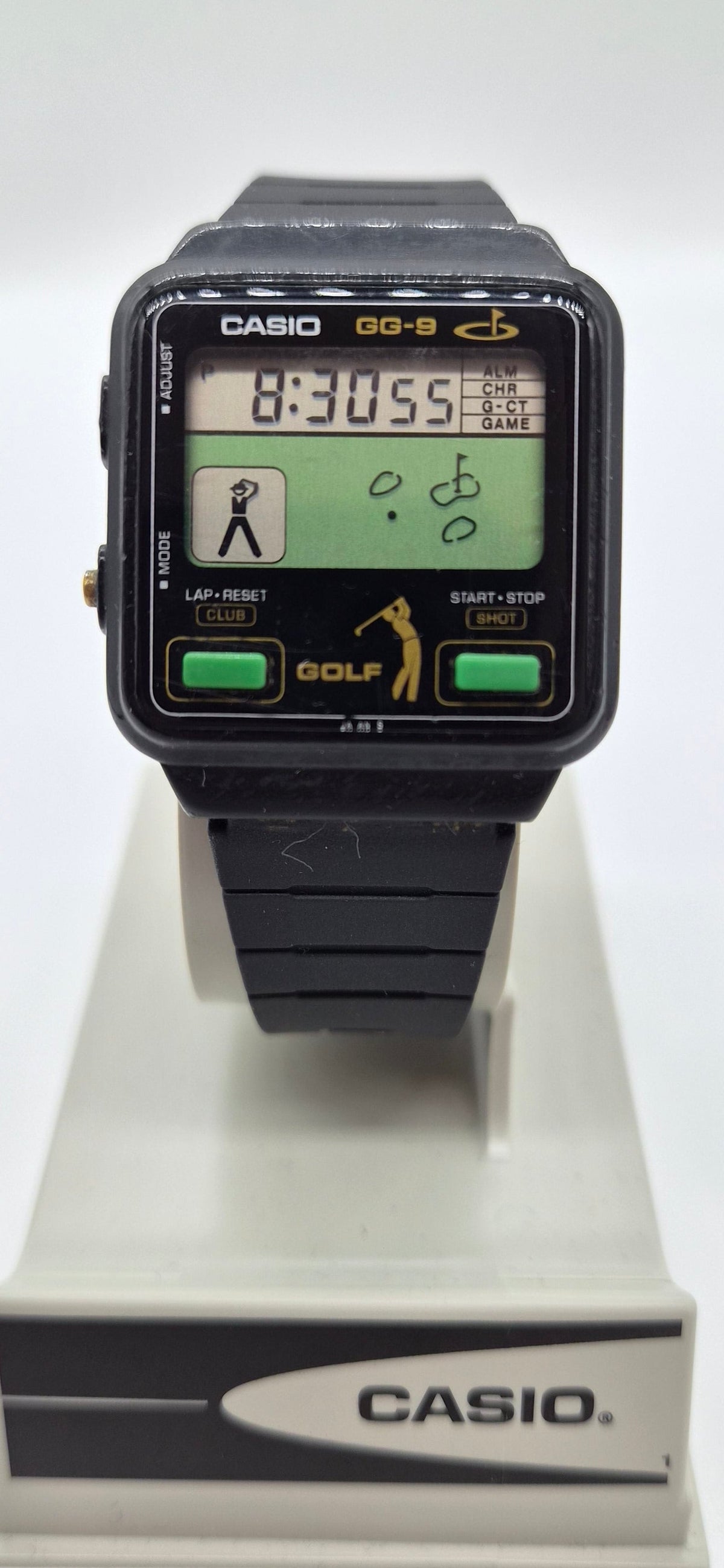 A Casio GQ-9 wristwatch with a black rectangular case, green LCD display, and black strap, displayed on a white Casio branded stand against a white background.